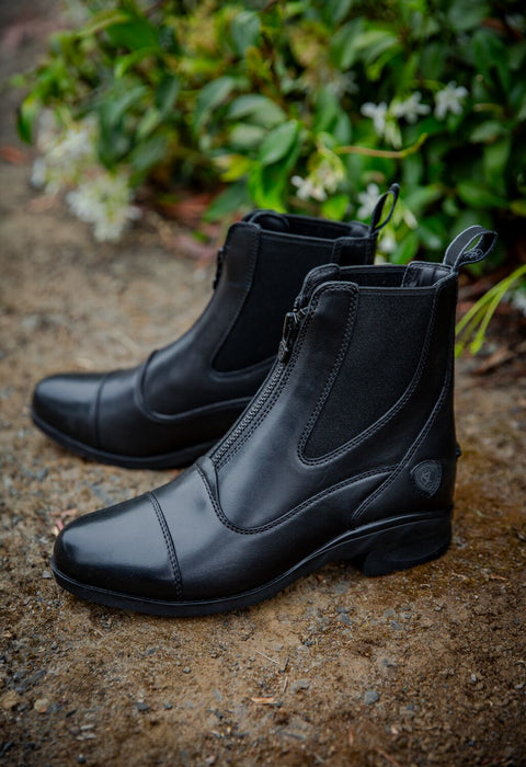 A pair of black leather equestrian riding boots with side zippers, displayed on a textured stone surface with green foliage in the background.