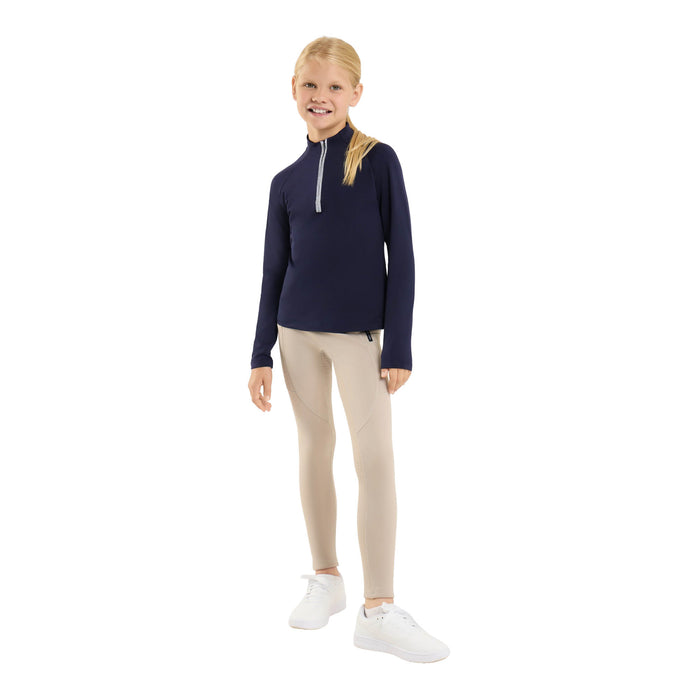 A young girl stands against a solid white background wearing a navy blue long-sleeve quarter-zip athletic top, beige riding breeches, and white sneakers.