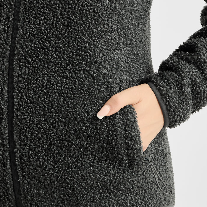 Close-up of a person wearing a dark charcoal gray sherpa fleece zip jacket, showing the textured fabric, a black zipper, and a hand tucked into a side pocket against a light gray background.