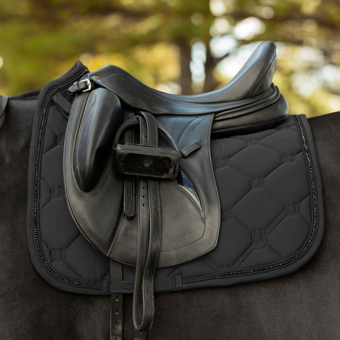 A close-up of a black leather horse saddle resting on a black quilted saddle pad, with the horse's dark coat and a blurred outdoor background of green and yellow foliage.