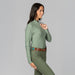A woman modeling a sage green long-sleeve athletic top with textured knit details on the chest, paired with olive green pants and a brown belt, against a solid gray studio background.