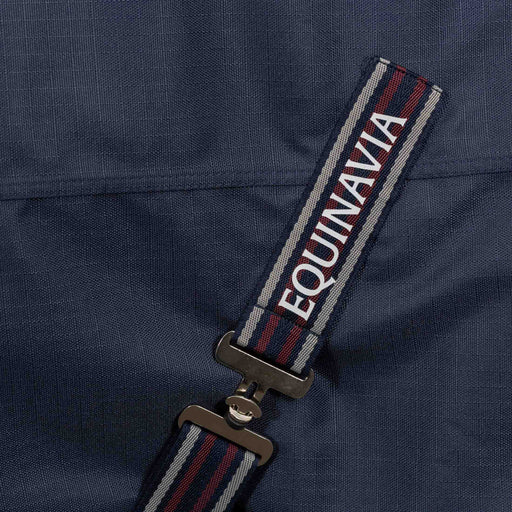 Close-up of a navy blue fabric surcingle strap with red and gray stripes and the brand name 'EQUINAVIA' in white, attached to a metal clip on a navy blue turnout blanket.
