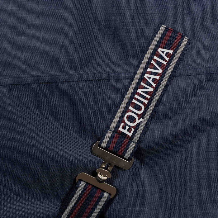 Close-up of a navy blue fabric surcingle strap with red and gray stripes and the brand name 'EQUINAVIA' in white, attached to a metal clip on a navy blue turnout blanket.