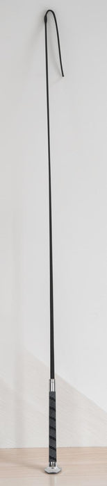 A long black riding dressage whip or whip with a curved hook-like tip, standing upright on a light wood floor against a plain white wall.