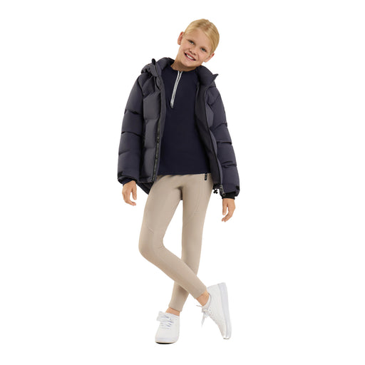 A young girl modeling a dark navy puffer jacket over a navy top, paired with beige leggings and white sneakers, on a solid white background.