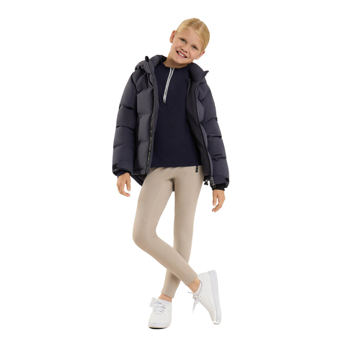 A young girl modeling a dark navy puffer jacket over a navy top, paired with beige leggings and white sneakers, on a solid white background.