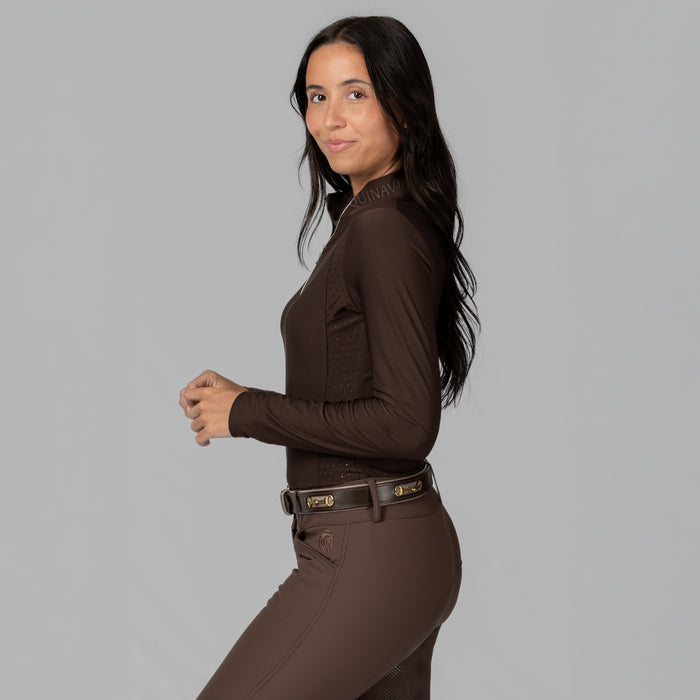 A woman modeling a dark brown long-sleeve equestrian shirt and matching brown riding pants against a solid gray studio background.