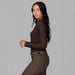 A woman modeling a dark brown long-sleeve equestrian shirt and matching brown riding pants against a solid gray studio background.