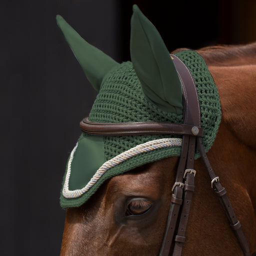 A close-up of a horse wearing a green knit fly veil with attached ear covers under a brown leather bridle, shown against a dark background.