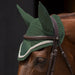 A close-up of a horse wearing a green knit fly veil with attached ear covers under a brown leather bridle, shown against a dark background.