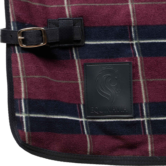 Close-up of a plaid fleece blanket with a burgundy, navy, and white pattern, featuring a black strap with a brass buckle and a black leather Equinavia brand patch.