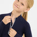 A close-up of a young girl wearing a navy blue long-sleeve athletic top with a silver-gray zipper, smiling and pulling the zipper with both hands against a clean white background.