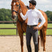 A man in equestrian attire, including a white long-sleeve shirt, dark grey riding breeches, and black riding boots, stands next to a chestnut horse in an outdoor riding arena.