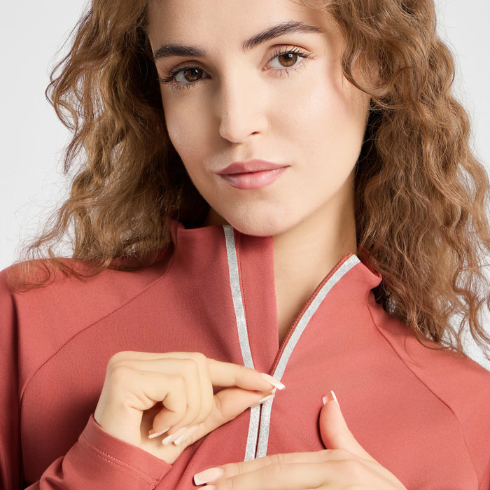 A close-up of a woman wearing a dusty rose athletic zip-up jacket with a silver-gray trim along the collar and zipper. She is looking directly at the camera with her hands near the front zipper.